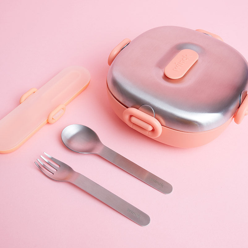 Winck stainless steel fork and spoon laid out next to a Peach bento lunch box with matching clasp detailing, all against a pink background, highlighting the brand's commitment to combining function with a playful color palette.