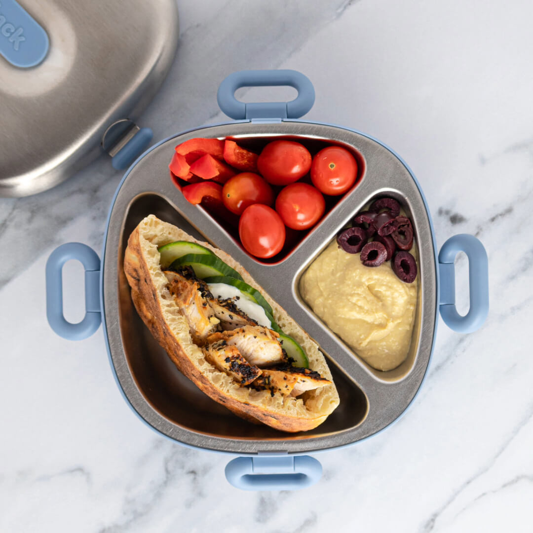 A Winck stainless steel bento box containing a Mediterranean-style lunch: a mini pita pocket filled with grilled chicken, cucumber slices, and tzatziki sauce, accompanied by cherry tomatoes, sliced red bell peppers, hummus, and kalamata olives.