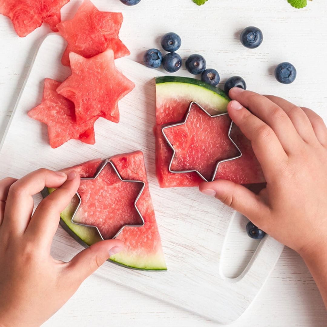 Hands of a child using star-shaped cookie cutters to cut out pieces from slices of watermelon, with blueberries and mint leaves scattered around on a white background.