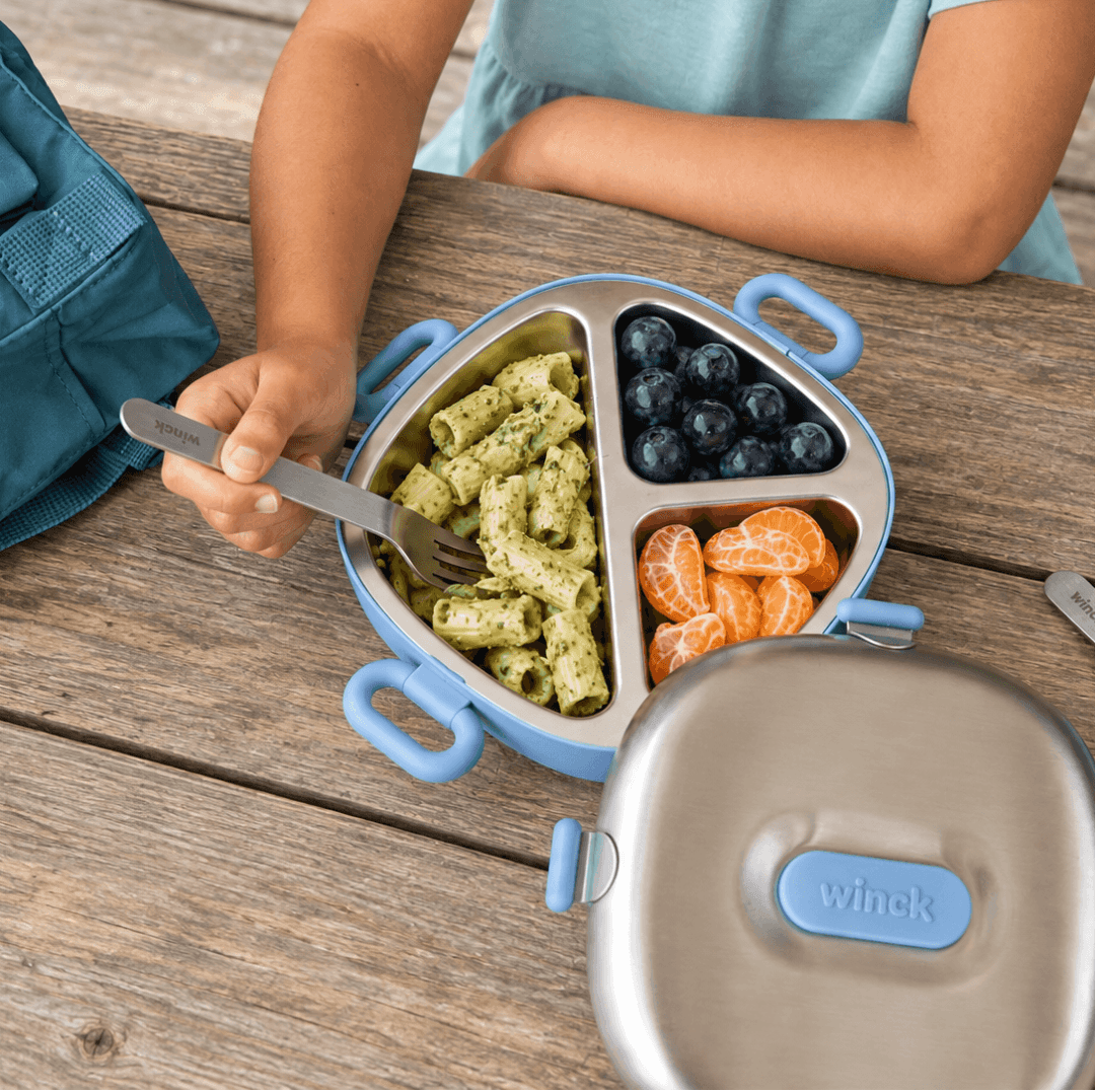 A child sitting at a wooden picnic table with a Winck three-compartment lunch container. The stainless steel container has light blue handles and compartments, filled with pasta, blueberries, and orange slices, with the matching lid in the foreground.