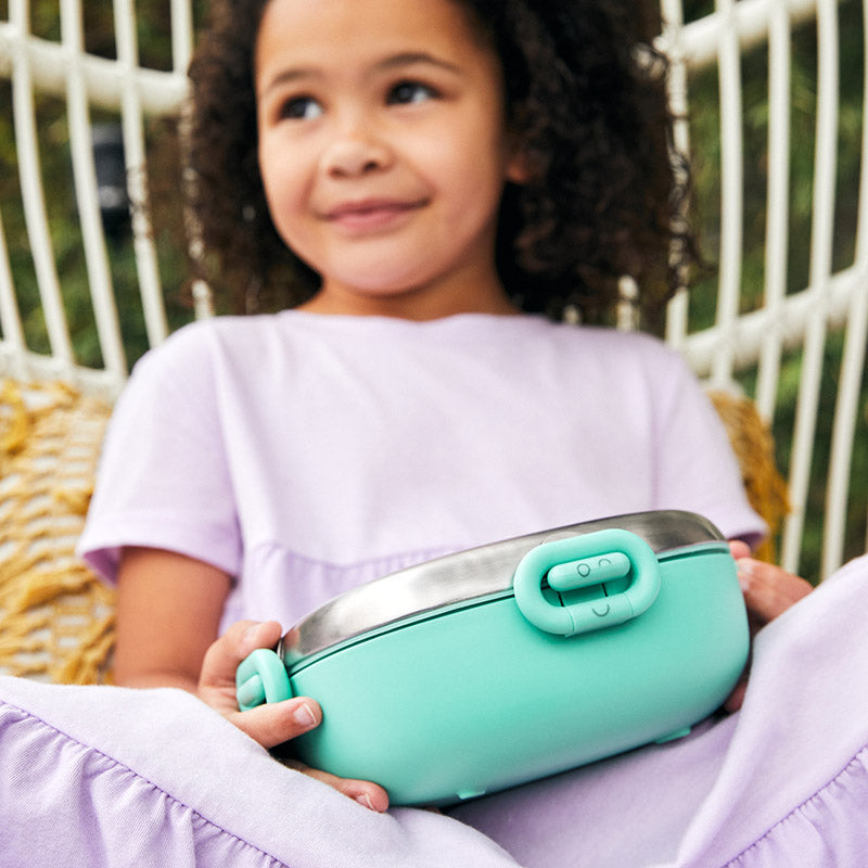 A young girl is smiling and sitting on a chair outdoors with a Winck Stainless Steel Bento Lunch Box for Kids in Pistachio on her lap.