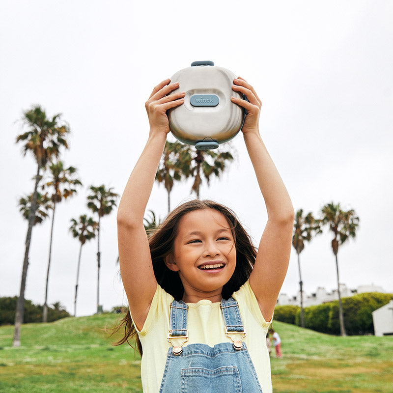 Joyful girl holding a Winck stainless steel bento lunch box above her head in an outdoor park, with a backdrop of tall palm trees, symbolizing fun and freedom with healthy on-the-go eating for kids.