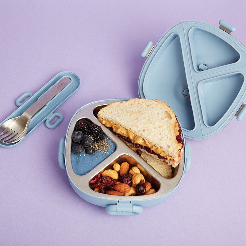 Stainless steel Winck bento lunch box on a purple background, filled with a hearty peanut butter and jelly sandwich, a mix of nuts and dried fruits, and fresh blueberries with chia seeds, beside matching utensil set, ready for a nutritious meal.