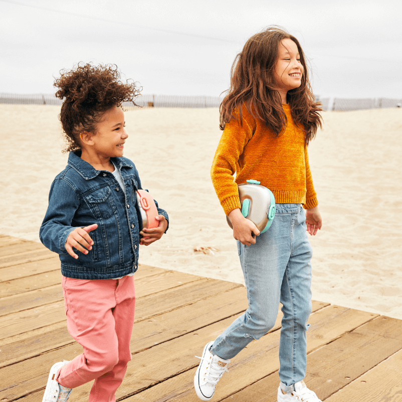 Two joyful children walking on a beach boardwalk, each carrying a Winck stainless steel bento lunch box, depicting a carefree lifestyle and the convenience of portable, healthy lunches for active kids.