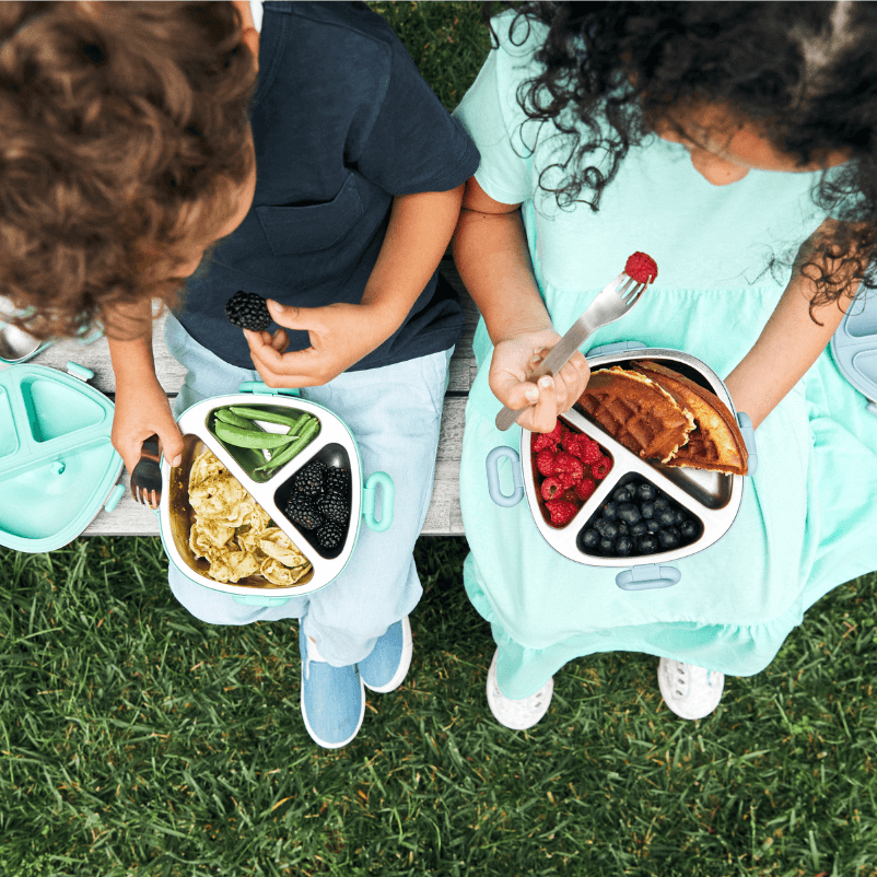 Overhead shot of two children seated in a park, each with an open bento box filled with a healthy variety of foods including pasta, berries, waffles, and snap peas, capturing a moment of outdoor dining and enjoyment.